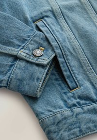 Világoskék farmerdzseki ujjával, kis fedőzsebbel, amelyen fémgomb és látható narancssárga varrás található.