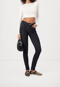 Jeans skinny de mezclilla oscura con cintura alta, combinados con una camiseta ajustada de manga larga blanca y bailarinas de estampado de leopardo. Bolso negro con asas.