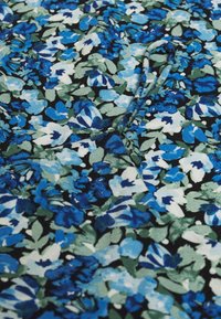 Blommönstrad tyg med nyanser av blått och grönt på en svart bakgrund, med en mjuk textur och en lätt glans. Synliga sömnadsdetaljer.