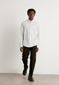 Hvit, langermet skjorte med knapper og lysegrå striper; paret med svarte bukser og mørke sneakers, frontalvisning. Enkelt design, uten mønstre.