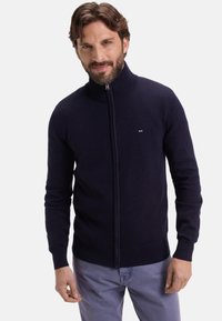 Homme avec une barbe portant un pull à fermeture éclair bleu marine et un pantalon gris clair, se tenant devant un fond blanc uni.