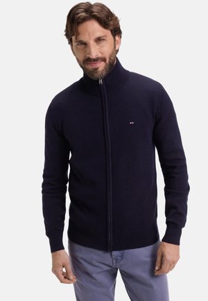 Homme avec une barbe portant un pull à fermeture éclair bleu marine et un pantalon gris clair, se tenant devant un fond blanc uni.