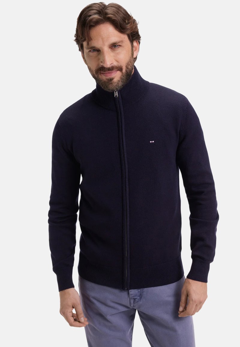 Homme avec une barbe portant un pull à fermeture éclair bleu marine et un pantalon gris clair, se tenant devant un fond blanc uni.