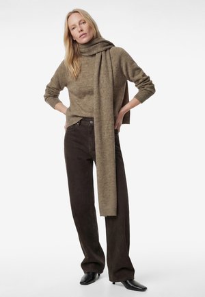 Frau mit langen blonden Haaren, die einen braunen Rollkragenpullover, einen passenden langen Schal, dunkelbraune weite Hosen und schwarze Stiefel trägt.