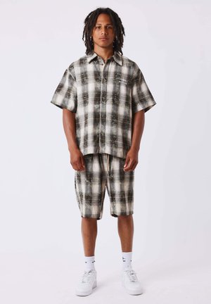 Jeune homme avec des dreadlocks aux épaules portant une chemise à manches courtes à carreaux assortie et un short avec des baskets blanches et des chaussettes.