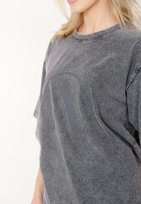 Szary, oversize T-shirt wykonany z miękkiego materiału, z dużym, wyblakłym nadrukiem litery "A" na środku. Krótkie rękawy i okrągły dekolt.