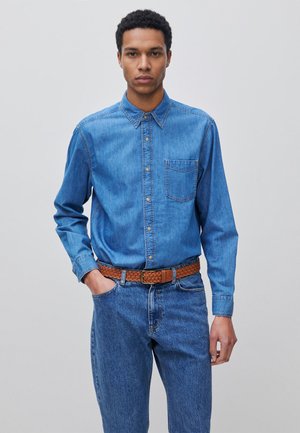 Jonge man draagt een blauw denim overhemd in een blauwe spijkerbroek met een bruine gevlochten riem, staand met één hand in zijn zak.