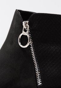 Stivaletto in suede nero con una cerniera visibile sul lato, dotato di un lucchetto argentato lucido e dettagli testurizzati vicino alla cerniera.