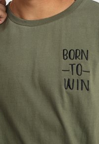 Camiseta de algodón verde oliva con cuello redondo, que presenta texto bordado en negro "NACIDO - PARA - GANAR" en la parte superior izquierda.