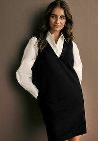 Robe noire sans manches avec un col en V, superposée sur une chemise blanche à boutons. Texture lisse, longueur genou, design minimaliste sans motifs.