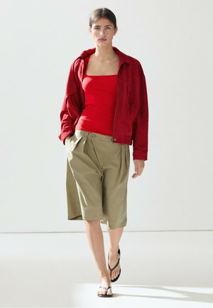 Donna che cammina indossando una giacca rossa sopra una canotta rossa, pantaloncini beige al ginocchio e sandali infradito neri su sfondo bianco.