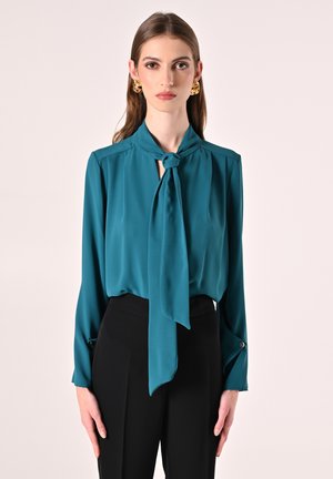 Blusa di seta verde acqua con dettaglio al collo annodato, maniche lunghe e bottoni ai polsini, abbinata a pantaloni neri a vita alta.