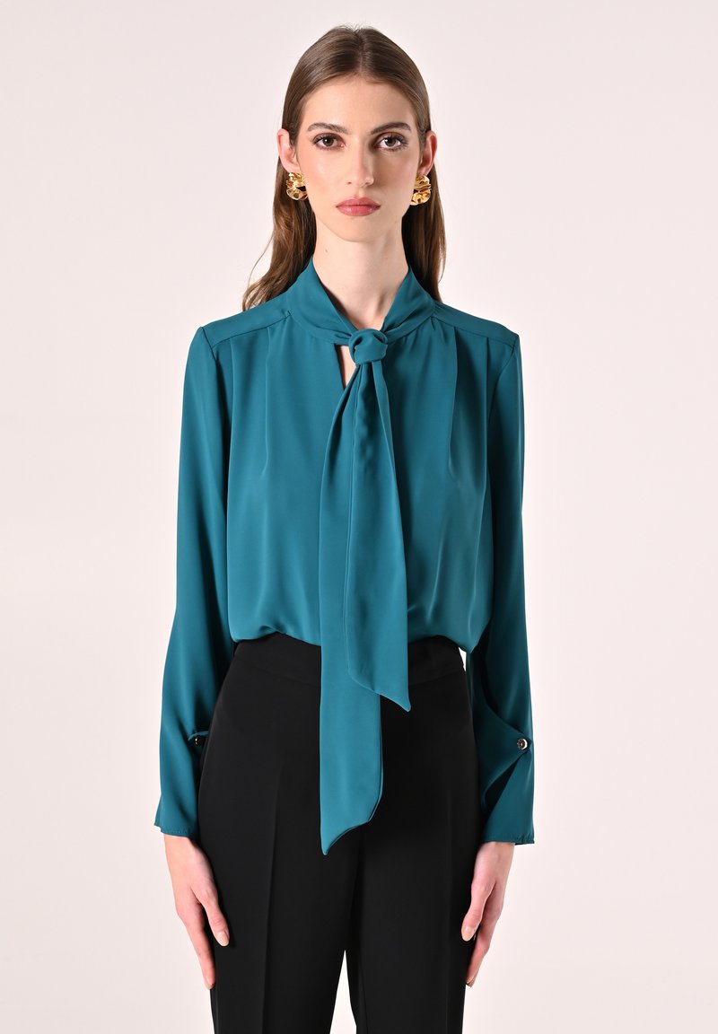 Blusa di seta verde acqua con dettaglio al collo annodato, maniche lunghe e bottoni ai polsini, abbinata a pantaloni neri a vita alta.
