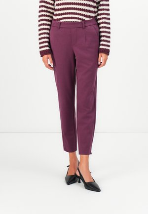 Femme portant un pantalon d'ourlet ajusté couleur prune, un pull en maille rayé, et des escarpins noirs à bride arrière, debout devant un fond blanc.