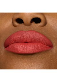 Matte koral læbestift på fyldige læber; glat tekstur og defineret form; rig farvekontrast mod mørkere hudtone. Nærbillede.