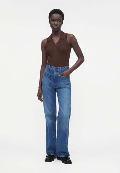 Γυναίκα στέκεται φορώντας αμάνικο καφέ ριμπ πουκάμισο με γιακά, τζιν παντελόνι με ψηλή μέση και μεγάλες εμπρόσθιες τσέπες και μαύρα μοκασίνια.