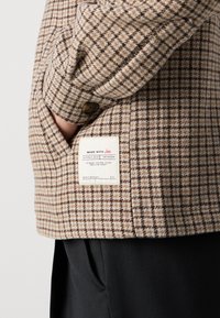 Chaqueta de cuadros marrón con un tejido texturizado, detalle de botones y una etiqueta con instrucciones de cuidado cosida en el lateral.