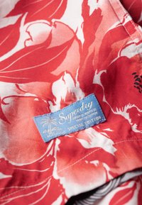 Rood bloemmotief stof met een blauw label met de tekst "Superdry Special Edition," versierd met palmboomgraphics en textuursteken.
