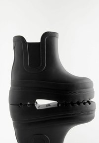 Botte de cheville en caoutchouc noir avec une finition texturée, des panneaux latéraux élastiques et une semelle épaisse présentant un design découpé distinctif.