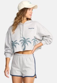 Vrouw in een lichtgrijze cropped hoodie met palmboomprint, bijpassende shorts met marineblauwe rand en een beige pet, kijkt opzij.