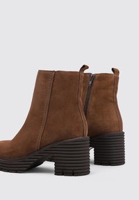 Bottines en cuir suédé marron avec un bout arrondi, une fermeture éclair sur le côté et des talons empilés cannelés. La texture est lisse avec des détails de couture subtils.