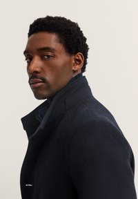 Marynarka w kolorze granatowym z wysokim kołnierzem, z teksturowanej tkaniny, z akcentem w postaci srebrnego zamka błyskawicznego. Nosi się ją na ciemnym golfie, co podkreśla dopasowany krój.