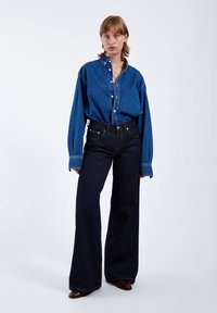 Outfit in denim composto da una camicia azzurra chiara, grande e con bottoni, e jeans a vita alta di un blu scuro, svasati, dotati di cinque tasche e cuciture a contrasto.