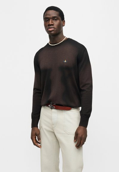 Άνδρας που φοράει μαύρο πουκάμισο με μακριά μανίκια και μικρό κεντητό λογότυπο, λευκό παντελόνι, καφέ ζώνη και κολιέ με πέρλες, στέκεται μπροστά σε λευκό φόντο.