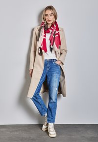 Beige lange jas, witte t-shirt, blauwe versleten jeans en een gestreepte rood en roze sjaal. Bruine en crèmekleurige sneakers met veters, staande tegen een grijze muur.