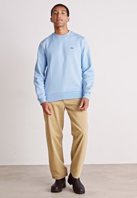 Sweatshirt bleu clair avec un col rond, des poignets et un ourlet côtelés. Associé à un pantalon beige et des bottines noires. Branding minimal visible.