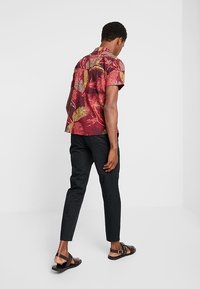 Camicia a maniche corte con sfondo rosso e motivi di foglie gialle e verdi, abbinata a pantaloni neri attillati e sandali marroni.