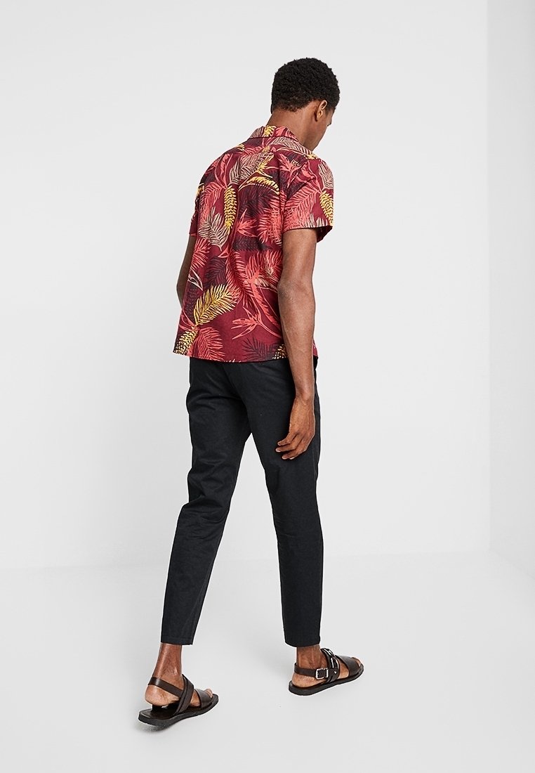 Camicia a maniche corte con sfondo rosso e motivi di foglie gialle e verdi, abbinata a pantaloni neri attillati e sandali marroni.