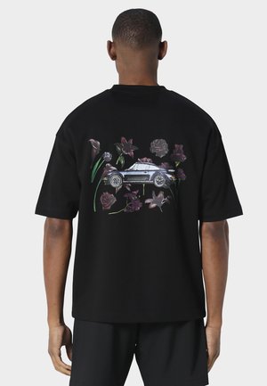 Person iført en sort T-shirt med et motiv af en klassisk bil omgivet af mørkerøde og lilla blomster på bagsiden.