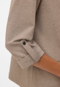 Jasnobrązowy długi rękaw z wywiniętym mankietem z czarnym guzikiem do zapięcia. Miękka, teksturowana tkanina.