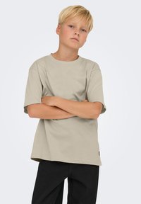 Camiseta de algodón beige con mangas cortas y escote redondo, con un corte holgado. Se lleva con pantalones negros ligeramente ajustados.