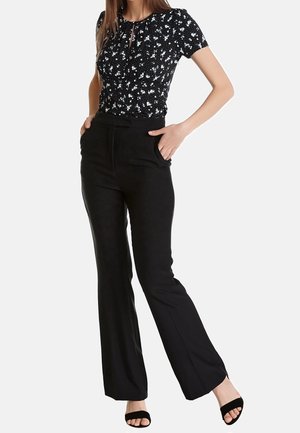 Mujer con blusa negra de flores y mangas cortas metida en pantalones anchos negros de talle alto, con las manos en los bolsillos y tacones negros abiertos en los dedos.