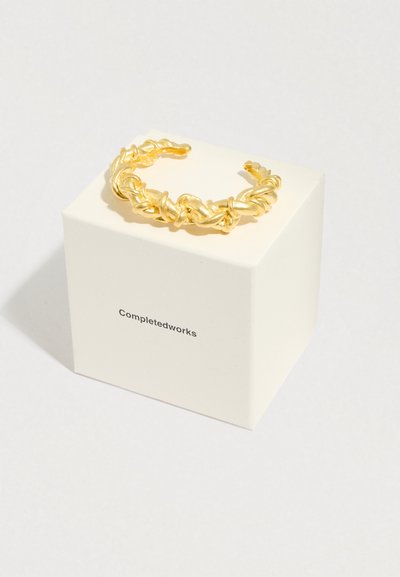 Pulseira de metal dourado com design torcido, repousando sobre uma caixa branca com o rótulo "Completedworks." Apresenta textura suave e extremidades abertas.