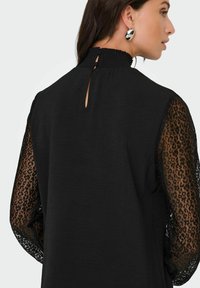 Zwarte top met een gestructureerd lijfje, doorschijnende lange mouwen met luipaardprint, hoge hals met geplooide details en een sluiting met een keyhole aan de achterkant.