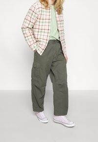 Rutigt jacka med gröna, rosa och bruna ränder, ljusgrön skjorta, olivgröna cargobyxor och ljusrosa sneakers. Avslappnad outfitdesign.
