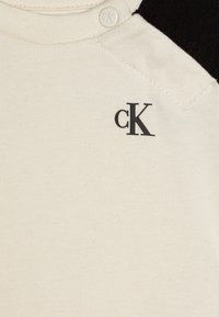 Ecru-kleurige katoenen t-shirt met zwarte raglanmouwen. Bevat een klein zwart 'cK' logo en een drukknoop dicht bij de hals.