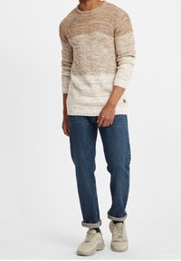 Texturerad stickad tröja i tan och krämfärgad gradient, rund halsringning, ihop med upplöpta mörka jeans och beiga sneakers.