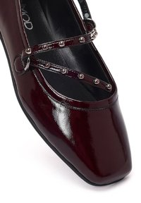 Chaussure en cuir verni bordeaux avec un bout carré et des sangles croisées décoratives ornées de clous métalliques le long des sangles. Texture de surface lisse.