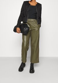 Giacca nera corta, pantaloni in similpelle verde, stivali neri combat, mentre tiene una pochette nera con texture. Design semplice e funzionale.