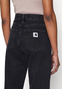 Svarte denimjeans med høy midje. Har en synlig sydd baklomme og en hvit logo-merke. Jevn tekstur, klassisk passform.