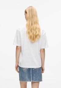 Mulher com cabelo loiro longo vestindo uma camisa branca de manga curta e uma saia de ganga azul desbotada, de pé de costas em um fundo branco.