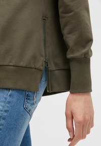 Olivengrüner Reißverschluss-Sweatshirt mit gerippten Bündchen, seitlichen Reißverschlusstaschen und glatter Textur, kombiniert mit abgewetzten hellblauen Jeans.