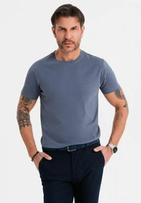 Camiseta de algodón gris con cuello redondo, mangas cortas y color sólido. Llevada con pantalones oscuros; el modelo tiene tatuajes en los brazos y un reloj en la muñeca.