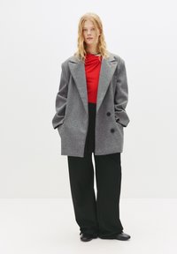 Cappotto in lana grigio oversize con due bottoni neri, indossato sopra un top rosso, abbinato a pantaloni neri larghi e scarpe nere. Sfondo luminoso e neutro.