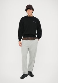 Felpa nera in cotone con testo "No Problemo", indossata sopra una maglietta marrone a fantasia. Pantaloni gris, sneakers nere e un cappellino marrone.