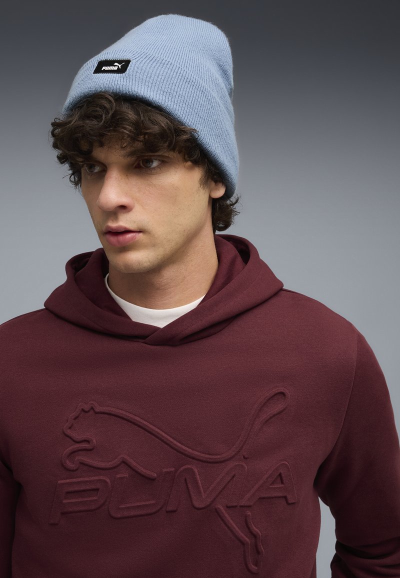 Bordo hoodie s utisnutim logotipom "PUMA", preko bijele majice. Svijetloplava rebrasta beanie dodatno kontrastira. Glatka tekstura, ležeran dizajn.